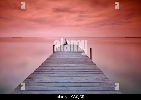 Méconnaissable personne assise sur le bord de la jetée en bois au coucher du soleil des couleurs rouge et violet Banque D'Images