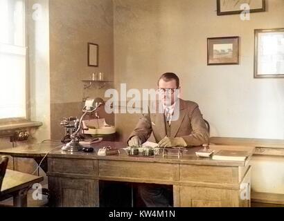 Un homme aux cheveux courts, des lunettes rondes, un costume conservateur et une expression faciale stern est assis derrière un grand bureau en bois dans son bureau, avec un petit lavabo, une encre buvard, deux cloches, une lampe de bureau, et d'un téléphone, de la Hongrie, 1929 visible. Numérisé à partir d'un processus de gélatine sur plaque de verre. Remarque : l'image a été colorisée numériquement à l'aide d'un processus moderne. Les couleurs peuvent ne pas être exacts à l'autre. Banque D'Images