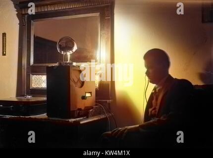 Un homme avec une expression sévère est assis dans une pièce sombre et écoute une grande radio qui se trouve sur un bureau, comme une petite lampe en haut de la radio se reflète sur un miroir au mur, illuminant la pièce d'ombres Stark, avec un porte-documents visibles sur le bureau, Hongrie, 1929. Numérisé à partir d'un processus de gélatine sur plaque de verre. Remarque : l'image a été colorisée numériquement à l'aide d'un processus moderne. Les couleurs peuvent ne pas être exacts à l'autre. Banque D'Images