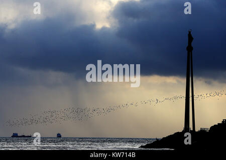 Une volée d'oiseaux voler au-delà de la "Notre Dame, Etoile de la mer' Memorial sur le mur Bull comme de sombres nuages en rouleau sur la baie de Dublin. Banque D'Images