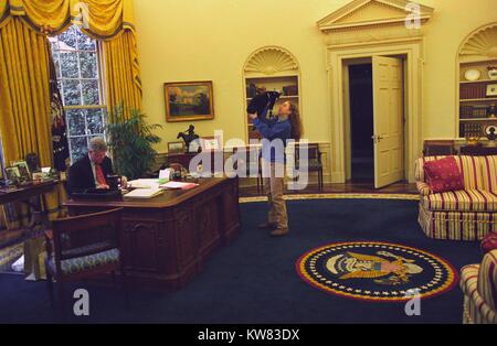 Le président Bill Clinton, portant costume et lunettes, travaille à son bureau dans le bureau ovale, tandis que la première fille Chelsea Clinton, vêtu d'un pantalon kaki et un pull bleu, se dresse en face de lui avec les bras tendus vers le haut, berçant le premier animal, des chaussettes le chat, avec pelage noir, visage blanc, et petit col, l'intérieur de la Maison Blanche, Washington, District de Columbia, le 24 décembre 1994. Banque D'Images