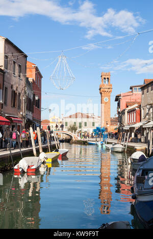 L'île de Murano, Venise, Italie, la Tour de l'horloge dans le Campo San Stefano reflétée dans Canal Vetrai avec bateaux colorés et touristes en hiver Banque D'Images