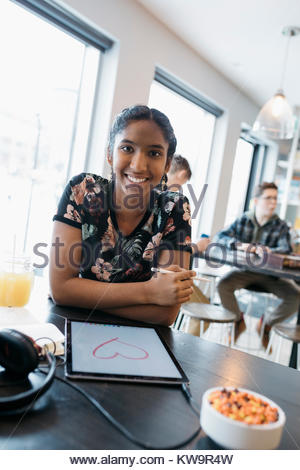 Portrait confiant Indian tween girl drawing heart-shape avec un stylet sur digital tablet in cafe
