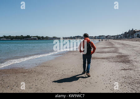 Jeune homme en veste rouge et un jean marche sur plage de Weymouth, Dorset, Angleterre sur une journée ensoleillée. Banque D'Images