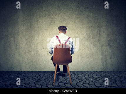 Vue arrière de l'homme assis sur une chaise à la recherche au mur gris en béton ayant des problèmes. Banque D'Images