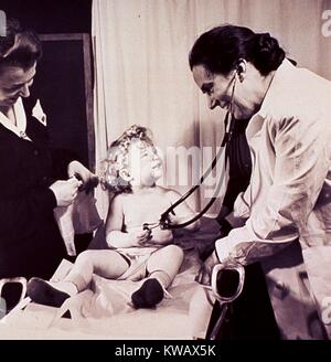 Un enfant, assis sur une table d'obstétrique, joue avec un stéthoscope du médecin au cours d'examen médical, 1950. La permission de la National Library of Medicine. () Banque D'Images