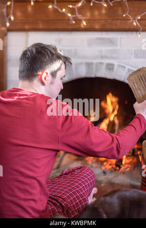 Jeune homme en pyjama assis sur le plancher à la maison avec un chien à partir d'un incendie sur un froid matin d'hiver Banque D'Images