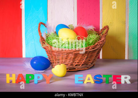 Panier en osier décoratif avec des oeufs peints et les mots joyeuses Pâques écrit avec des lettres en bois multicolore et d'une clôture à l'arrière-plan coloré. Banque D'Images