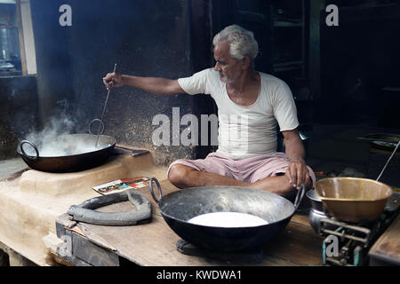 Scène de rue à Pushkar, inde Senior homme assis les jambes croisées sur le plancher de son atelier tandis que la cuisson dans une grande marmite, Rajasthan, Inde. Banque D'Images