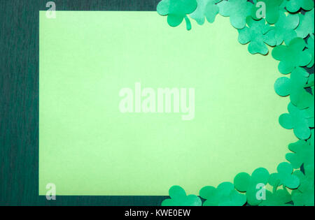 Saint Patrick carte de souhaits design avec beaucoup de trèfles papier entourant une feuille de papier jaune vide, affichée sur un fond de bois vert foncé. Banque D'Images