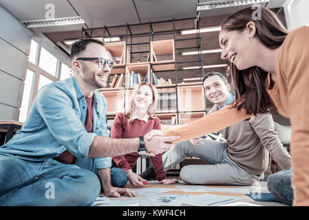 Relation d'affaires. Les jeunes employés satisfaits occupés assis sur le plancher près de leurs collègues shaking hands and smiling. Banque D'Images