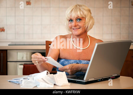 Femme d'âge moyen heureux assis dans sa cuisine à la table avec un ordinateur portable et un contrôle sur les prix sur les reçus du supermarché Banque D'Images