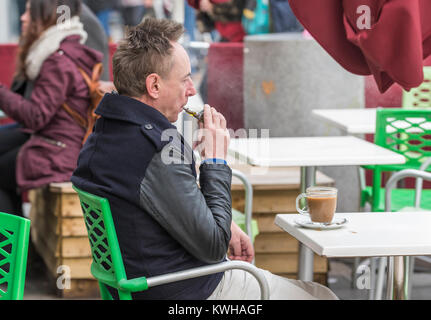 Homme caucasien assis dans un café à l'extérieur en hiver sur une cigarette électronique en Angleterre, au Royaume-Uni. Banque D'Images
