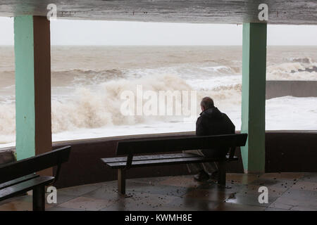 Hastings, East Sussex, UK. 4e janvier 2018. La marée a venir et la mer fait rage créant de grandes vagues se briser sur la jetée de Hastings et de la promenade. La vitesse du vent ont ramassé et des rafales de plus de 45 mph sont attendus plus tard cet après-midi. Le Met Office ont émis une alerte météo jaune de Hastings et le sud-est de l'Angleterre. Un homme assis sur un banc en bouteille Alley par Sidney peu donne sur la mer à la rage des vagues. Crédit photo : Paul Lawrenson/ Alamy Live News Banque D'Images