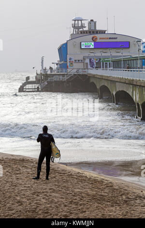 Bournemouth, Dorset, England UK. 4 janvier, 2018. Météo France : surfer debout sur la plage près de la jetée à la mer comme les surfeurs profiter du surf sur un jour de vent à la plage de Bournemouth, que les surfeurs profiter de la vent et de grosses vagues. Banque D'Images