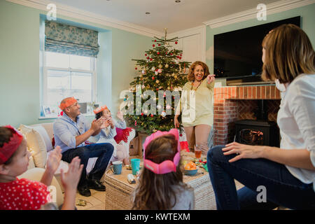 Famille jouent aux charades au moment de Noël dans le salon de leur maison. C'est au tour de la grand-mère et tout le monde rit et applaudit. Banque D'Images