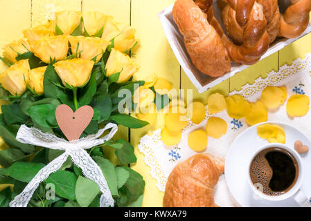 Repas du matin avec les produits de pâtisserie, une tasse de café aromatique et un gros bouquet de roses jaunes et les pétales, placé sur une table en bois jaune. Banque D'Images