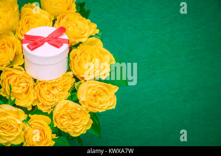 Magnifique bouquet de roses jaunes et un mignon petit cadeau blanc attaché avec du ruban rouge et bow au milieu, sur un fond de bois vert foncé. Banque D'Images