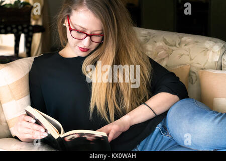 Une jeune blonde femme lisant un livre portant des lunettes, couché dans un canapé à la maison, le jour de la lumière naturelle. Banque D'Images