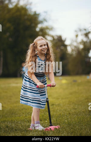 Jolie blonde enfant souriant sur scooter portant tenue d'été joue dans park Banque D'Images