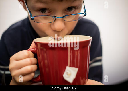 Un jeune garçon boit dans une tasse de thé dans une tasse rouge. Banque D'Images