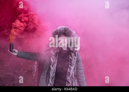 Jeune homme de race blanche dans un parka d'hiver vert posant avec une bombe fumigène rouge sur une sombre journée d'hiver. Tartu, Estonie. Banque D'Images