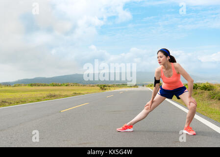 Young pretty female jogger portant des vêtements de sport avec appareil standing on country road stretching jambes réchauffer corps prêt à former d'entraînement. Banque D'Images