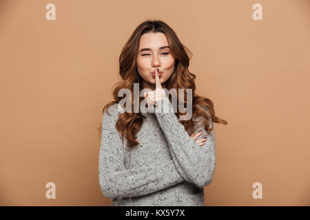 Close-up portrait of funny fille brune clignote un œil tout en montrant le silence, geste isolé sur fond beige Banque D'Images