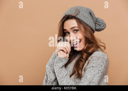 Photo en gros plan de cute brunette woman in hat chandail gris et réchauffement de la mains et looking at camera, isolé sur fond beige Banque D'Images