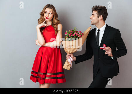 Portrait d'un homme joyeux qui se propose d'une jeune fille insatisfaite avec des fleurs et une bague de fiançailles sur fond de mur gris Banque D'Images