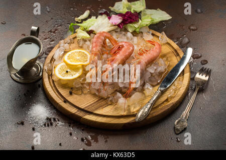 Grandes Crevettes tiger ou langostinos avec citron sur la glace sur planche à découper en bois. Cutler, de sauce et de laitue sur fond sombre. La cuisine méditerranéenne Banque D'Images