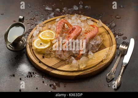 Grandes Crevettes tiger ou langostinos avec citron sur la glace sur planche à découper en bois. Cutler, sauce sur fond sombre. La cuisine méditerranéenne Banque D'Images