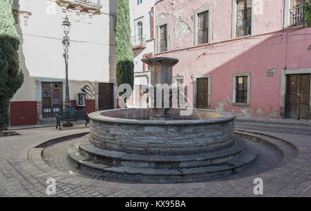 La fontaine en pierre dans la Plaza del Ropero, avec un banc en fer vert, certains bâtiments environnants et pavés, à Guanajuato, Mexique Banque D'Images