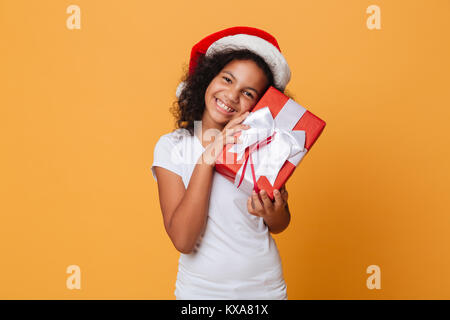 Portrait d'une jeune fille africaine peu satisfaits vêtus de christmas hat holding present fort et riant plus isolé sur fond orange Banque D'Images