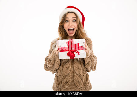 Choqué jeune femme habillé en pull-over chaud wearing christmas hat standing isolated over white wall background. Appareil photo à la holding gift box surpri Banque D'Images