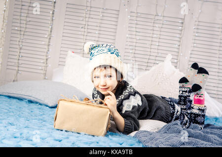 Petite fille l'enfant assis en pyjama et un chapeau sur le lit avec guirlande d'ampoules avec boîtes cadeaux enveloppés dans un papier coloré décoré de Banque D'Images