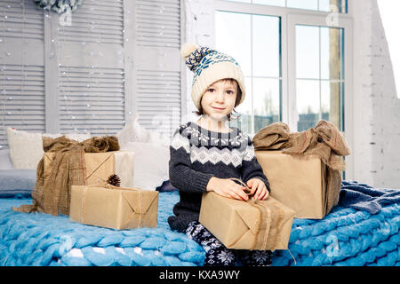 Petite fille l'enfant assis en pyjama et un chapeau sur le lit avec guirlande d'ampoules avec boîtes cadeaux enveloppés dans un papier coloré décoré de Banque D'Images