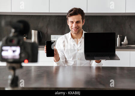 Beau jeune homme de filmer son blog vidéo épisode sur de nouveaux appareils de haute technologie tout en montrant l'écran vide téléphone mobile et ordinateur portable à l'intérieur Banque D'Images