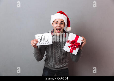 Image of happy man wearing christmas santa hat isolés sur mur gris holding paper avec vente texte et boîte-cadeau. À la caméra. Banque D'Images
