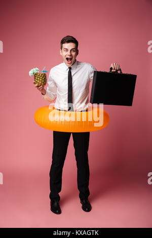 Photo de heureux heureux jeune homme avec bague en caoutchouc isolés holding suitcase et cocktail. À la caméra. Banque D'Images
