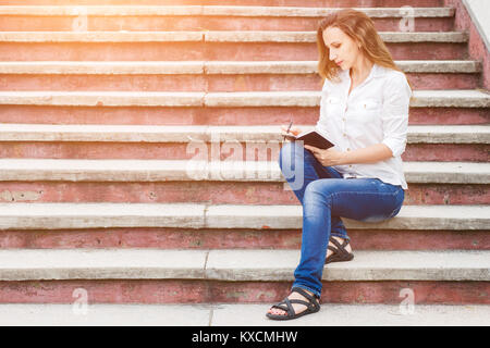Jeune femme assise sur les escaliers et prendre des notes dans l'ordinateur portable Banque D'Images