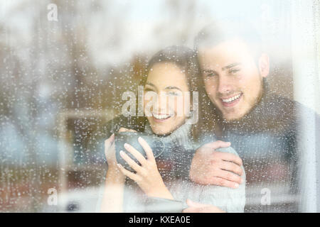 Vue avant, portrait d'un couple positif à l'extérieur par une fenêtre dans un jour pluvieux de l'hiver à la maison Banque D'Images