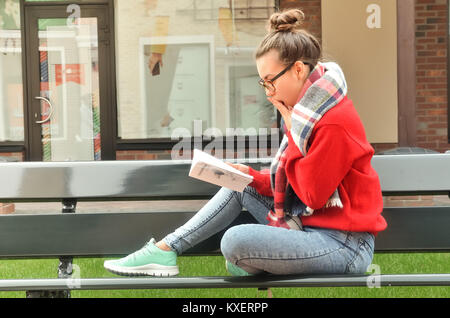 Une jolie fille asiatique portant des lunettes est assis sur un banc et lit un livre. Elle a été surprise et couvert sa bouche avec sa main. Derrière le photographe. Banque D'Images