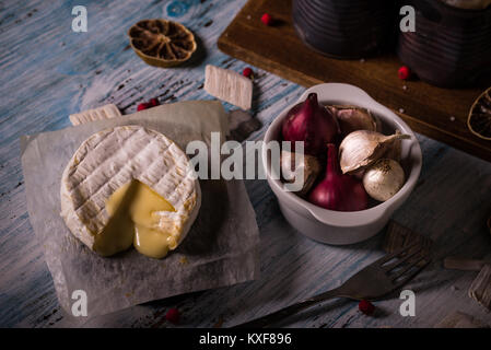 Photo horizontale de camembert fondu chaud sur feuille de papier avec la cuvette pleine d'ail et d'oignons et de vieux bidons vintage bleu porté sur planche de bois. Dri Banque D'Images