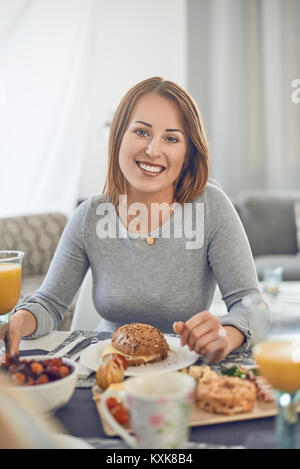 Happy attractive woman enjoying un sandwich pour le petit déjeuner assis à une table dressée avec de la nourriture smiling at the camera Banque D'Images