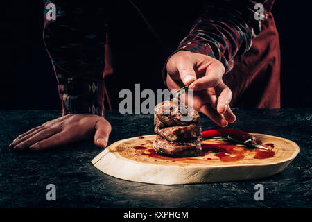 Close-up vue partielle de chef putting rosemary sur les steaks grillés avec sauce piment et sur planche de bois Banque D'Images