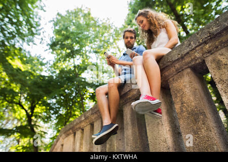 Jeune couple avec les smartphones en ville assis sur mur de béton. Banque D'Images