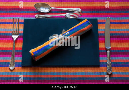 Couverts sur une serviette colorée sur un paillasson en ardoise noire - mise en place de table avec couteau, fourchettes et cuillère au-dessus de la tête Banque D'Images