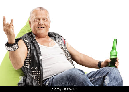 Vieux punk avec une bouteille de bière assis sur un pouf poire faisant un geste de la main de roche isolé sur fond blanc Banque D'Images