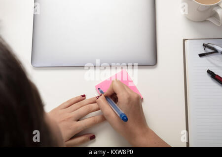 Femme écrivant une note adhésive rappel pour elle-même comme elle est assise à un bureau de travail avec un ordinateur portable et le bloc-notes avec stylo et verres Banque D'Images